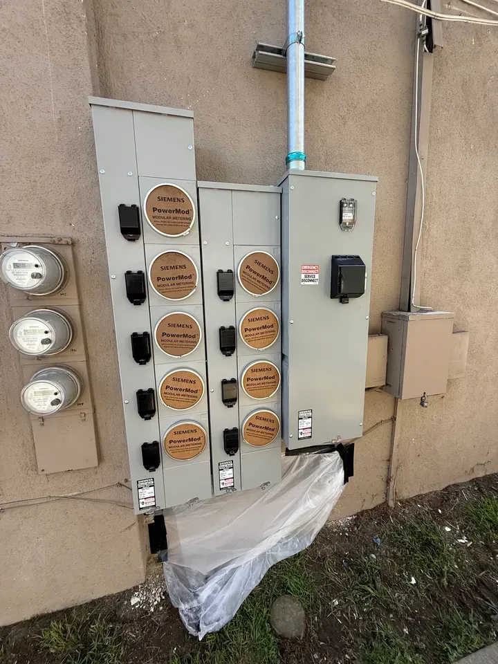 Outdoor electrical meter and service panels mounted on a tan wall with plastic sheeting below
