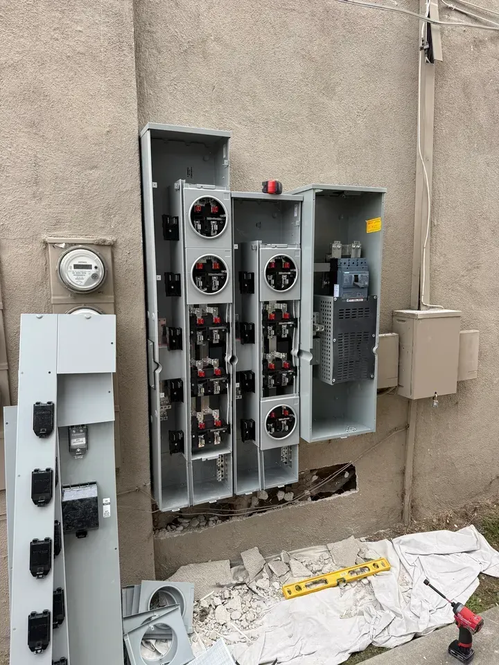 Open electrical panels on a beige exterior wall, with broken debris and tools on the ground.
