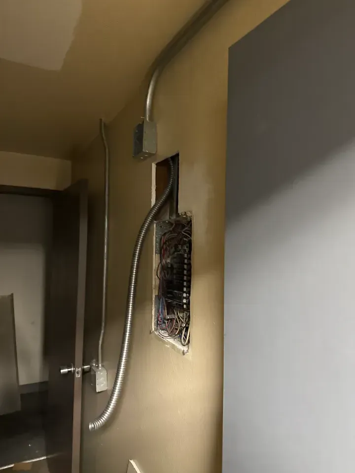Open electrical panel with exposed wiring on a beige wall in a dim hallway.