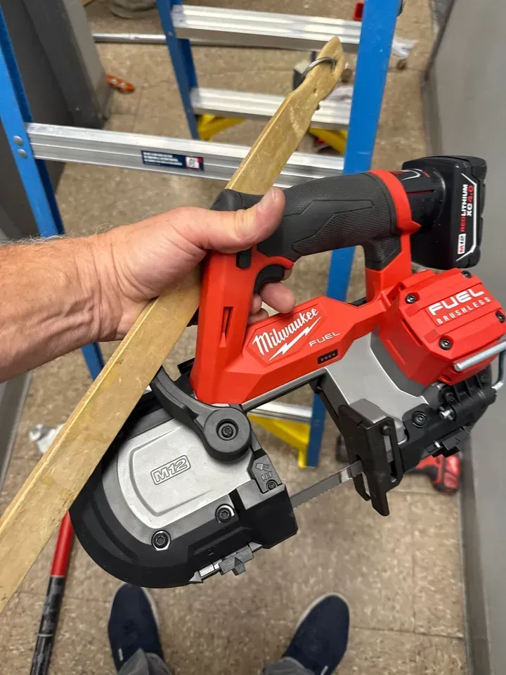 Hand holding a red Milwaukee cordless band saw in a workshop, with a ladder in the background