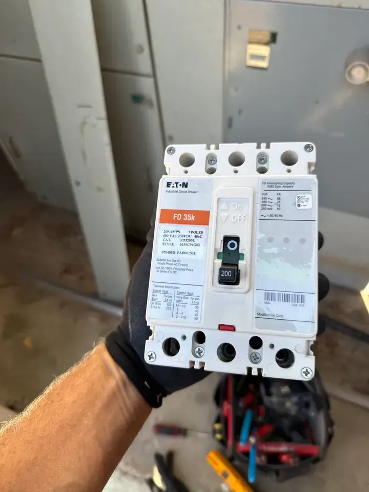 Hand holding a white electrical breaker panel in a utility room, with tools blurred in the background