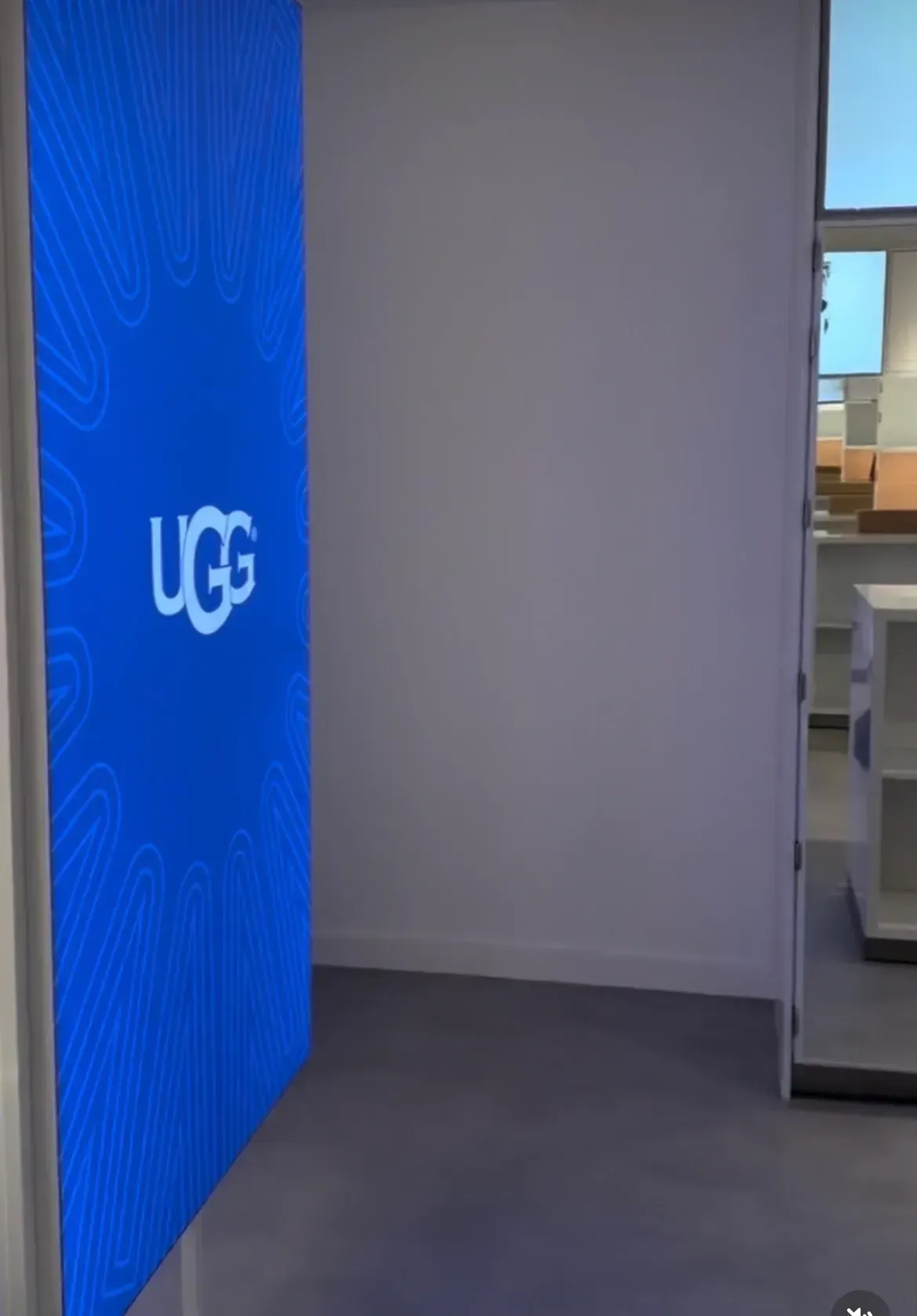 Blue UGG storefront display beside a white wall and doorway in a mall-like interior.