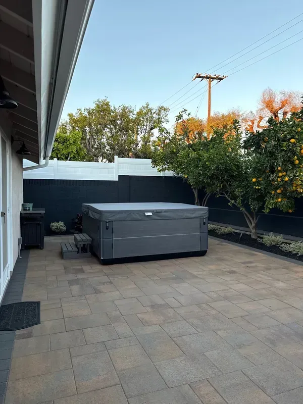 Patio with a gray hot tub beside a house, white fence, trees, and a paved stone floor.