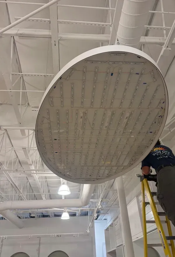 Worker on a yellow ladder inspecting a large round ceiling light in a white industrial interior
