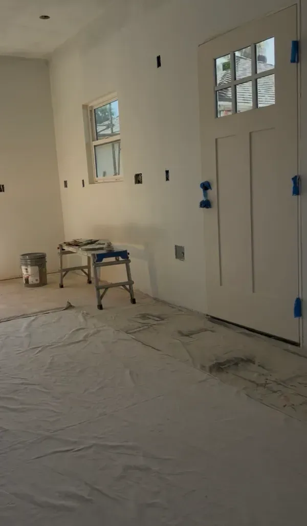 White interior room under construction with door, windows, a chair, and tools on a covered floor.