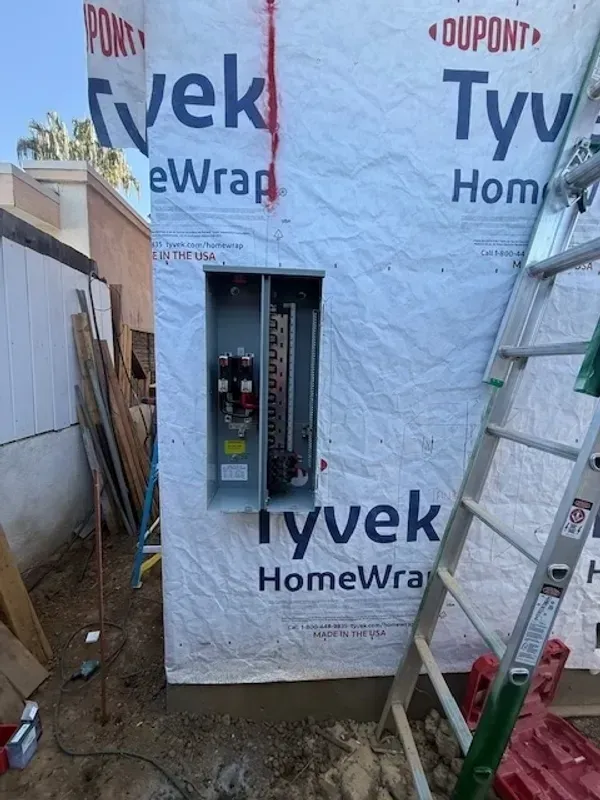 Breaker panel installed on Tyvek-wrapped exterior wall, with ladder beside it.