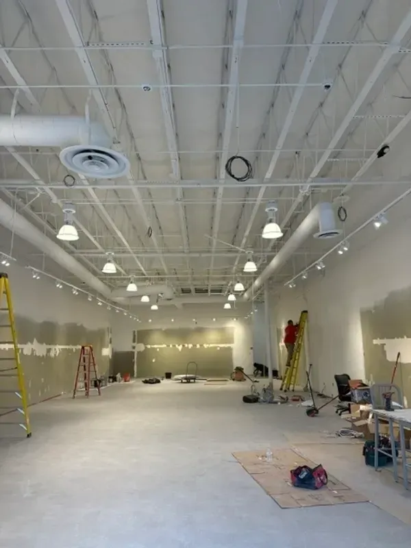 Empty warehouse-style room with high white ceiling, exposed lights, ladders, and scattered tools during renovation