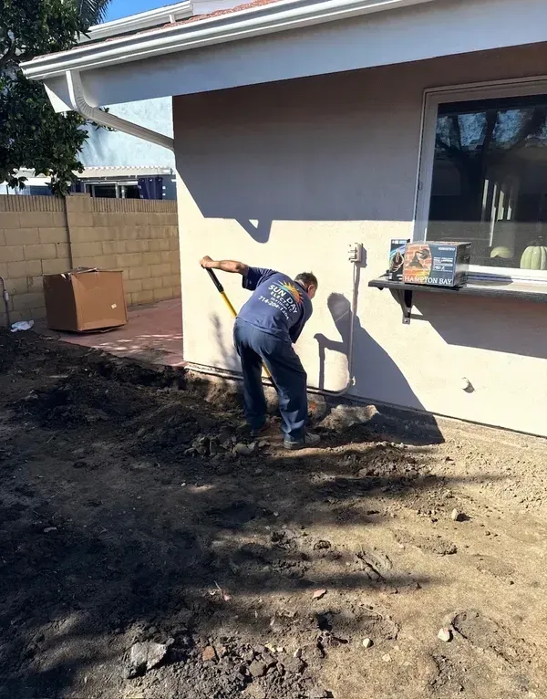 Worker shoveling dirt beside a house wall in a sunlit backyard