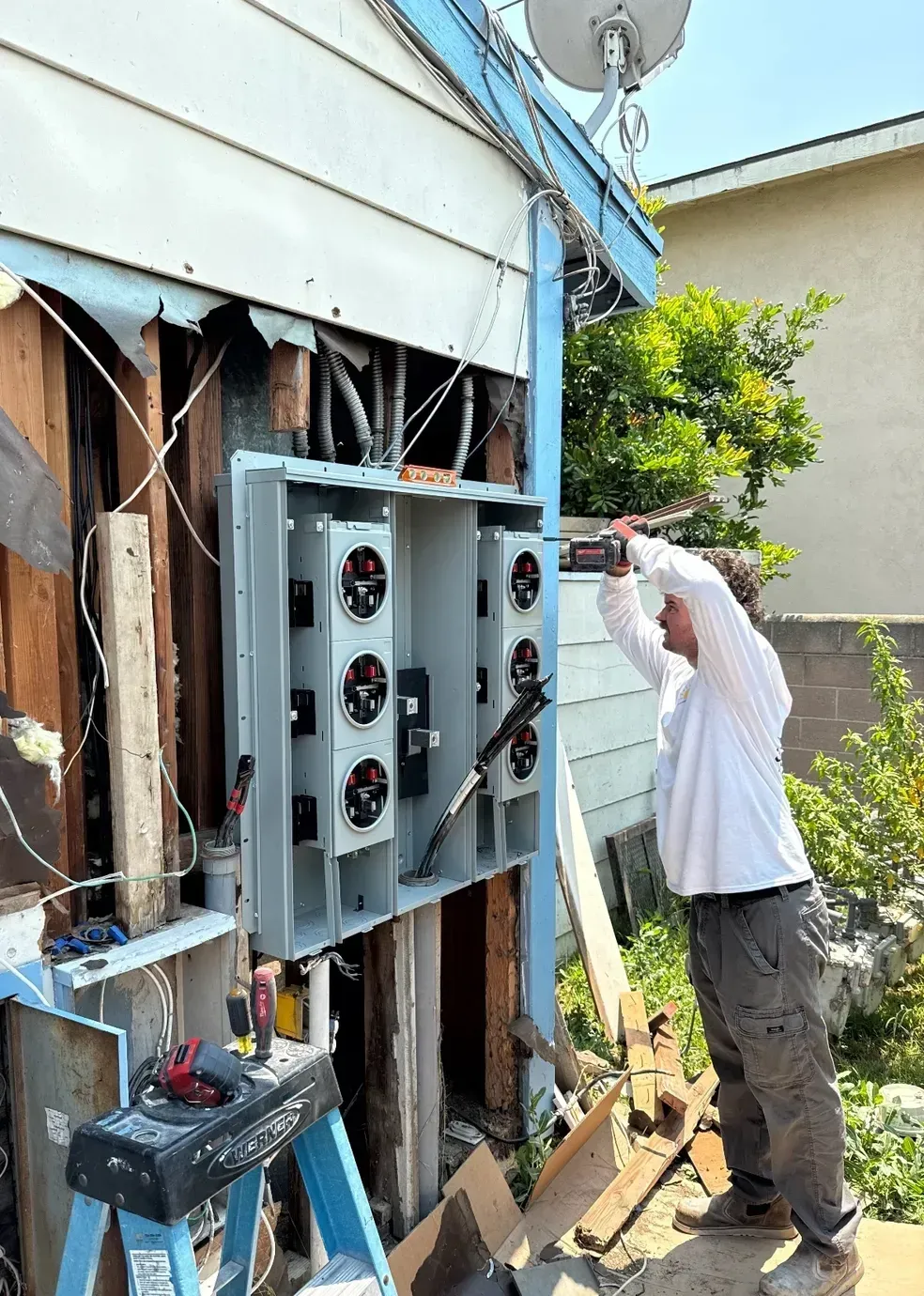 Worker inspecting electrical panels on the side of a house, with exposed wiring and tools nearby