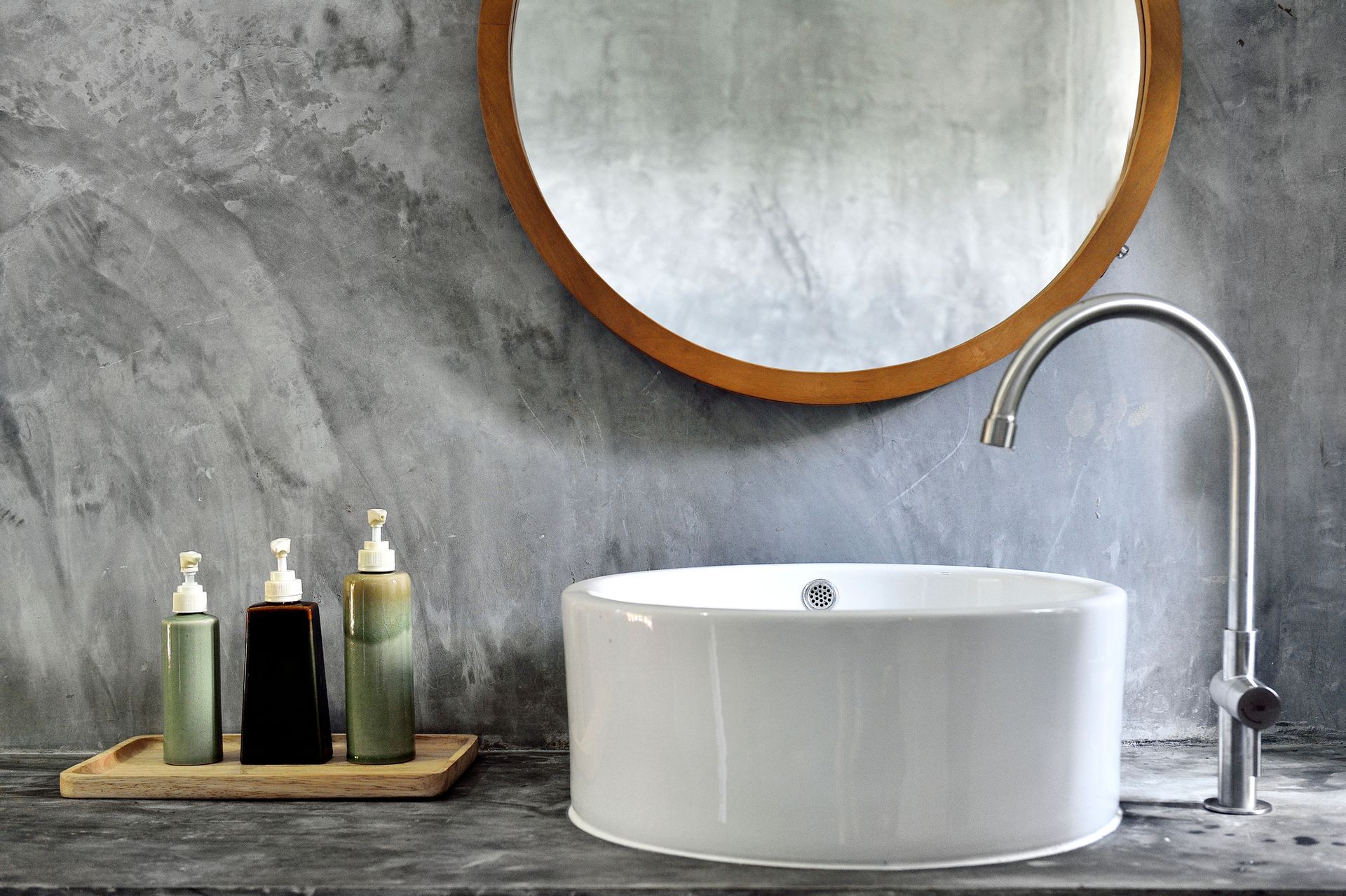 Lavandino del bagno con specchio rotondo, parete grigia e articoli da toeletta su un vassoio di legno.