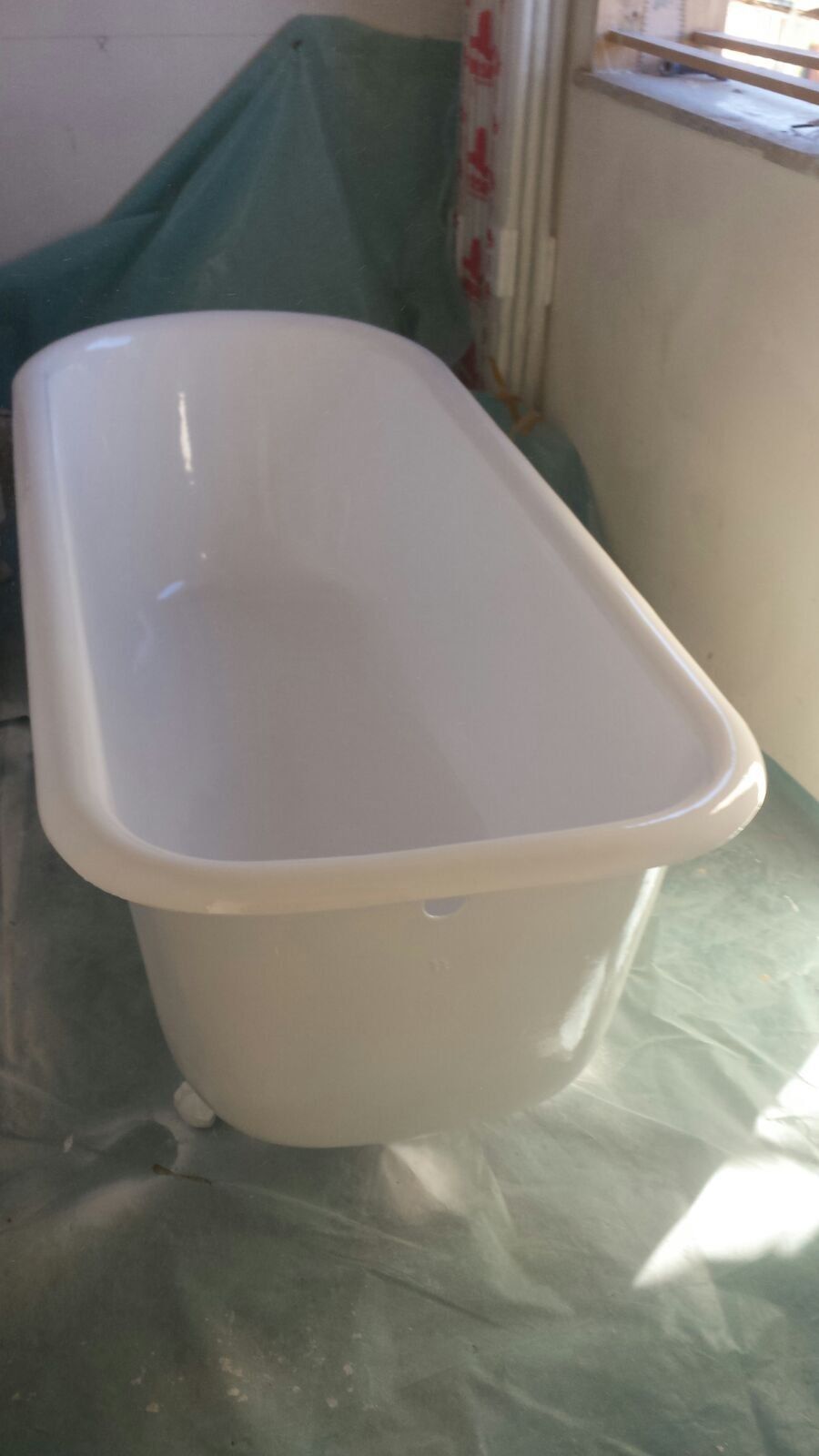 Vasca da bagno rettangolare bianca, appoggiata su un telo di plastica, vicino a una finestra.