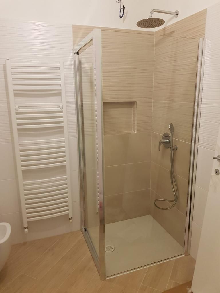 Bagno con doccia in vetro, piastrelle beige, portasciugamani e pavimento in legno chiaro.