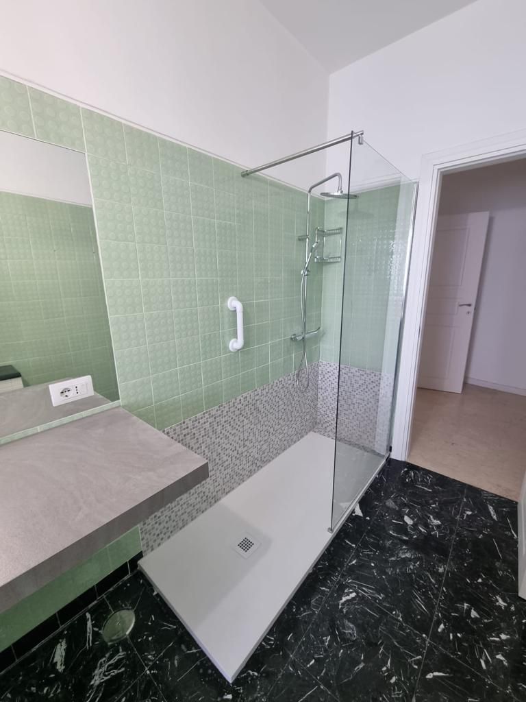 Bagno con pareti piastrellate verdi, box doccia in vetro e piano di lavoro grigio. Pavimento in marmo nero.