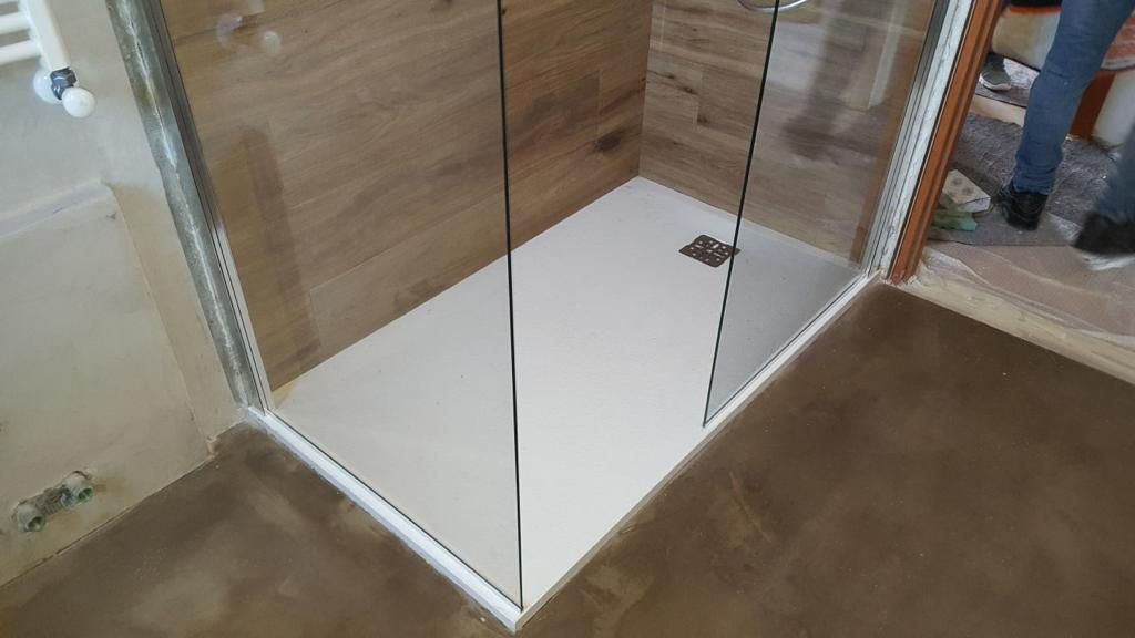 Box doccia con porte in vetro, base bianca, parete posteriore in legno e pavimento in cemento rifinito.