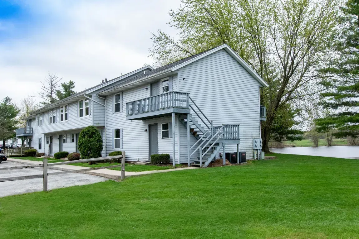 Two-story white apartment building with balconies and stairs beside a green lawn and parking area.