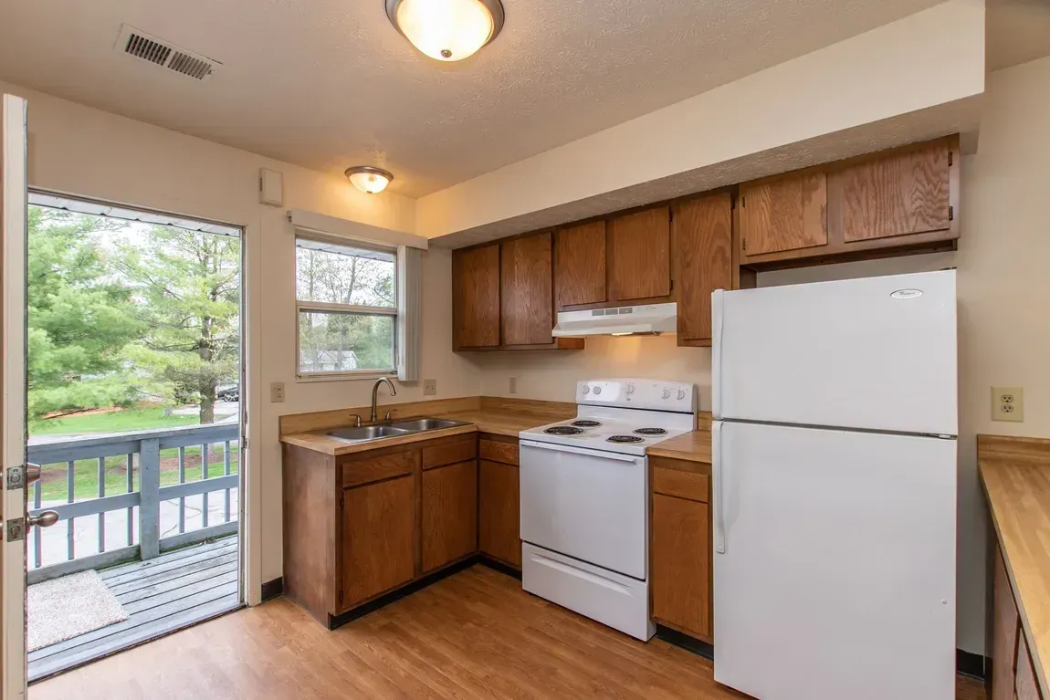 Small kitchen with wood cabinets, white stove and fridge, and a window over the sink.