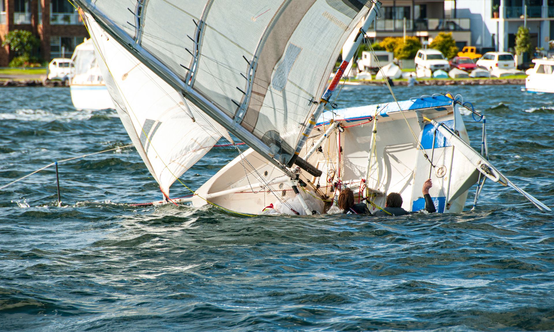 Small sailboat heeled over in choppy water, crew working on deck near a marina.