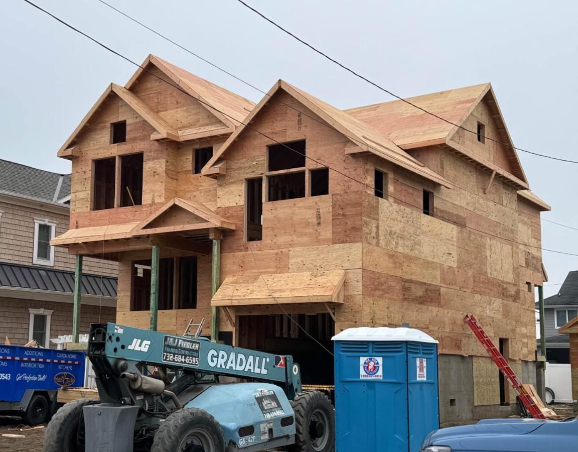 Construction site showing a two-story wood-frame house, a blue portable toilet, and a Gradall telehandler.