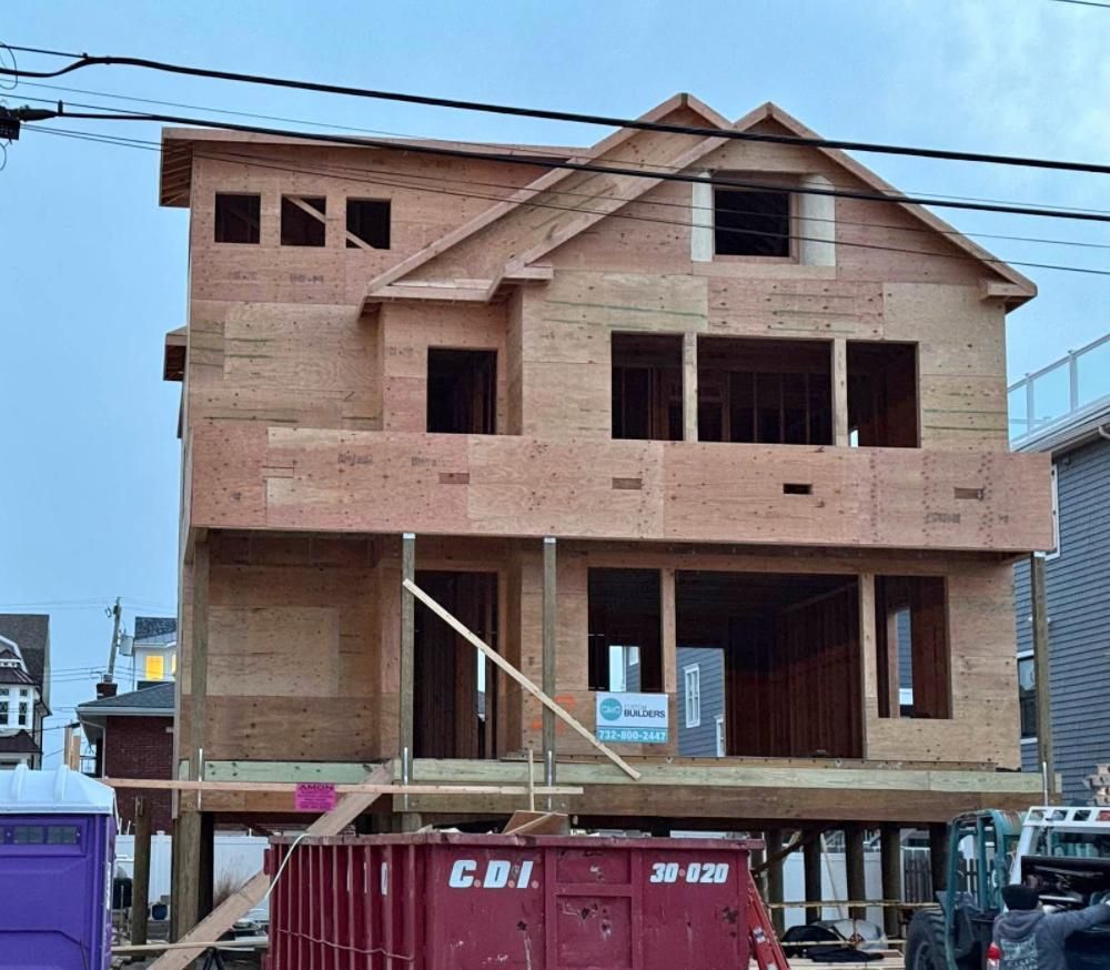 A three-story house under construction with exposed plywood framing, a red dumpster in the foreground, and utility lines.