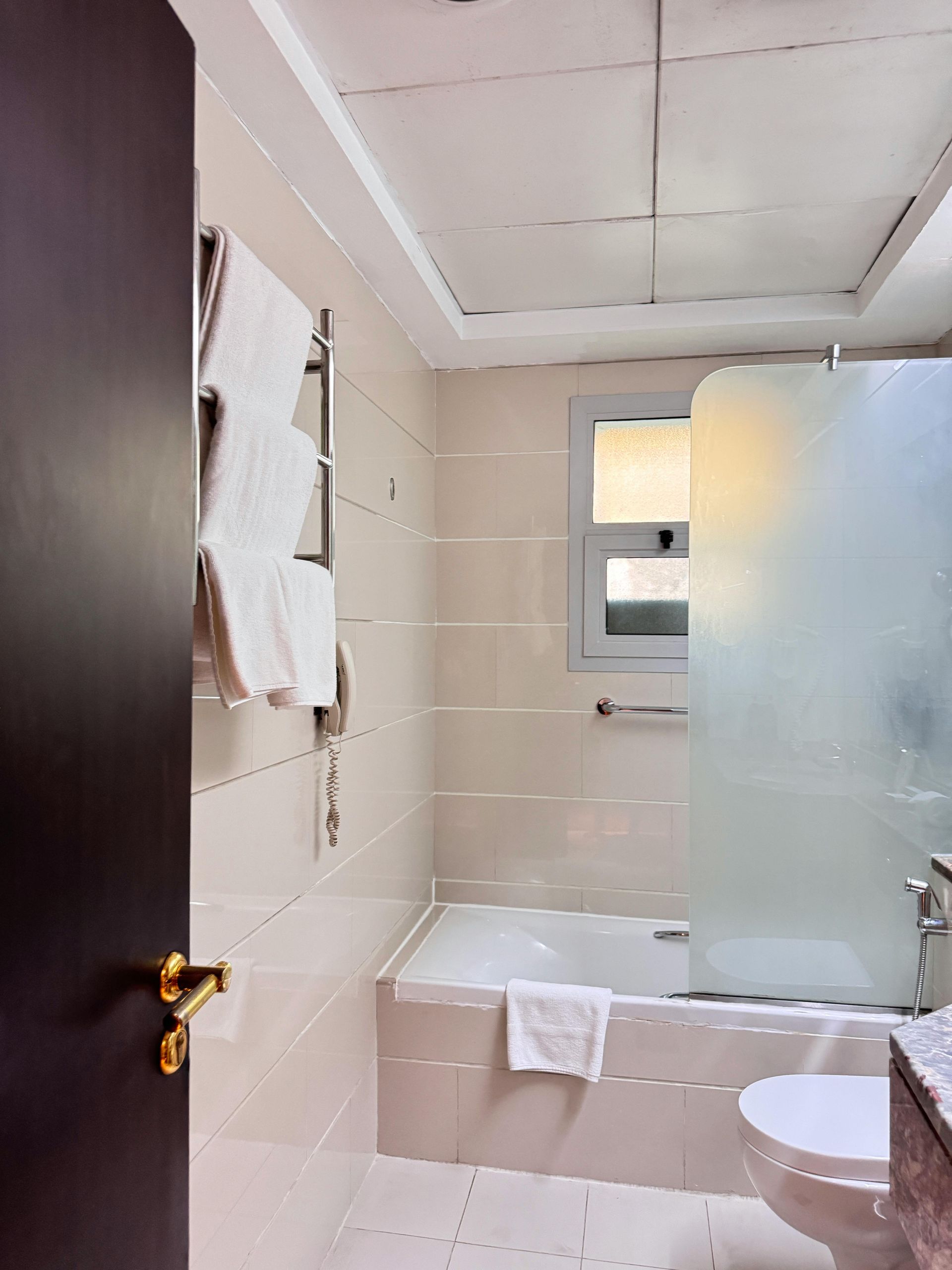 Small beige-tiled bathroom with a frosted shower door, sink, toilet, and towels hanging on the wall.