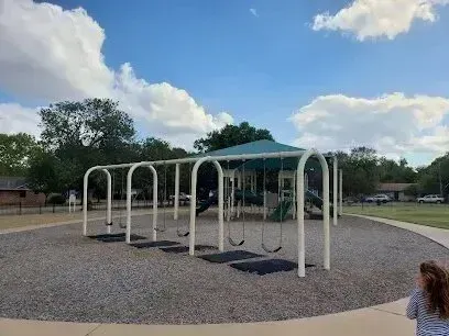 Burleson Electrician - A sunny playground with a set of empty swings on gravel, surrounded by trees. A child walks nearby, under a clear blue sky with fluffy clouds.