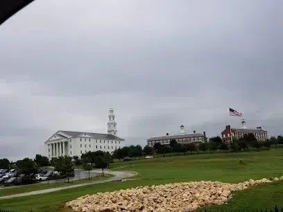 Burleson Electrician - Colonial-style white buildings with a prominent clock tower and an American flag stand atop a grassy hill under an overcast sky.
