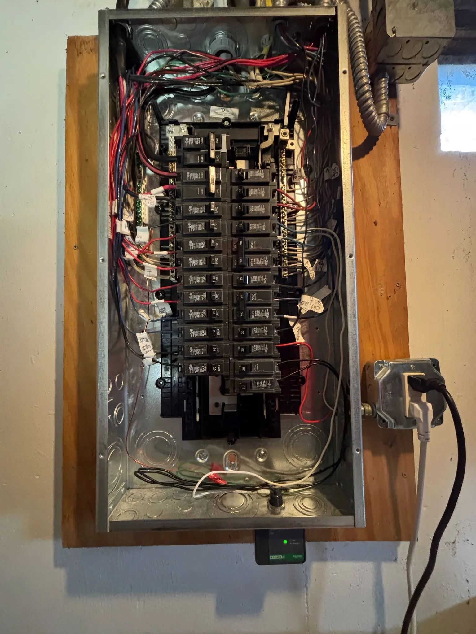Open electrical breaker panel with many wires, mounted in a wooden frame, with a corded device on the right.