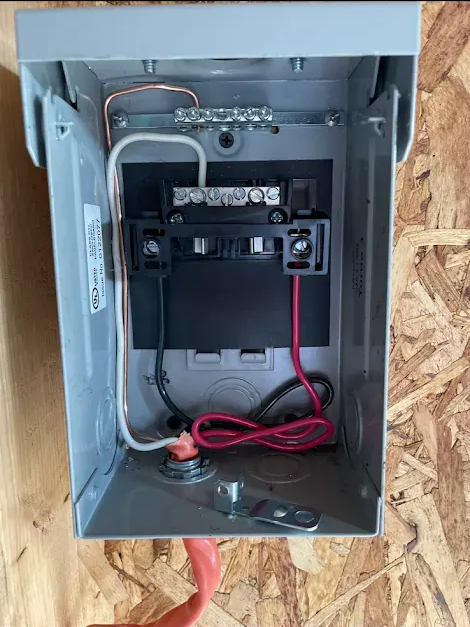 An open electrical disconnect box mounted on a wooden wall, showing internal wiring connections for power and ground.