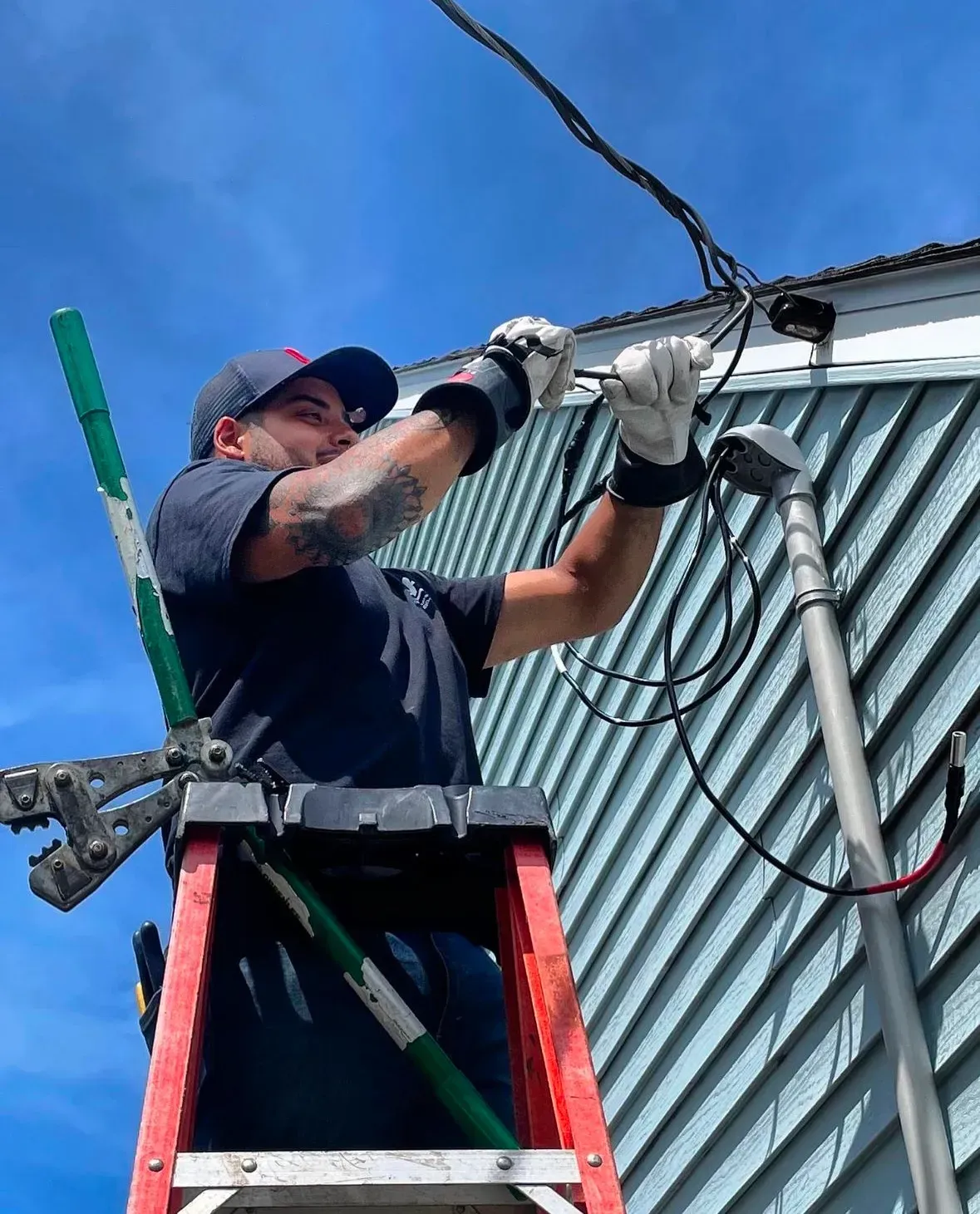 Lineman on a ladder, working on electrical wires near a blue house. Sunny, clear sky.