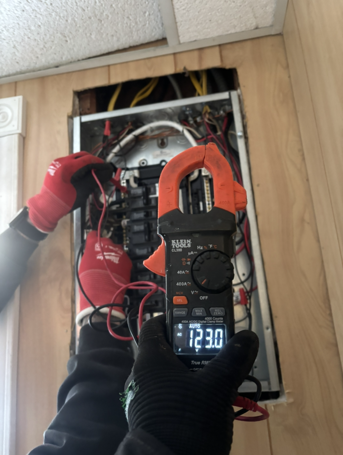 Hands testing exposed electrical panel with two multimeters and red/orange clamps