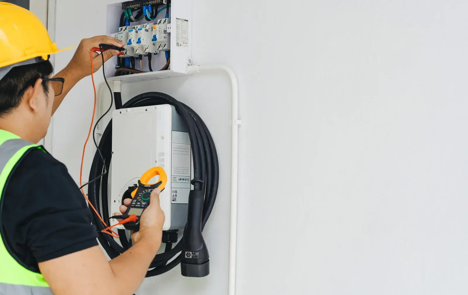 Electrician in yellow helmet tests electrical panel on white wall.