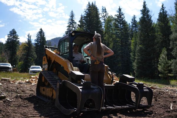 Two people on a yellow skid steer near a gate and barn in a rural yard