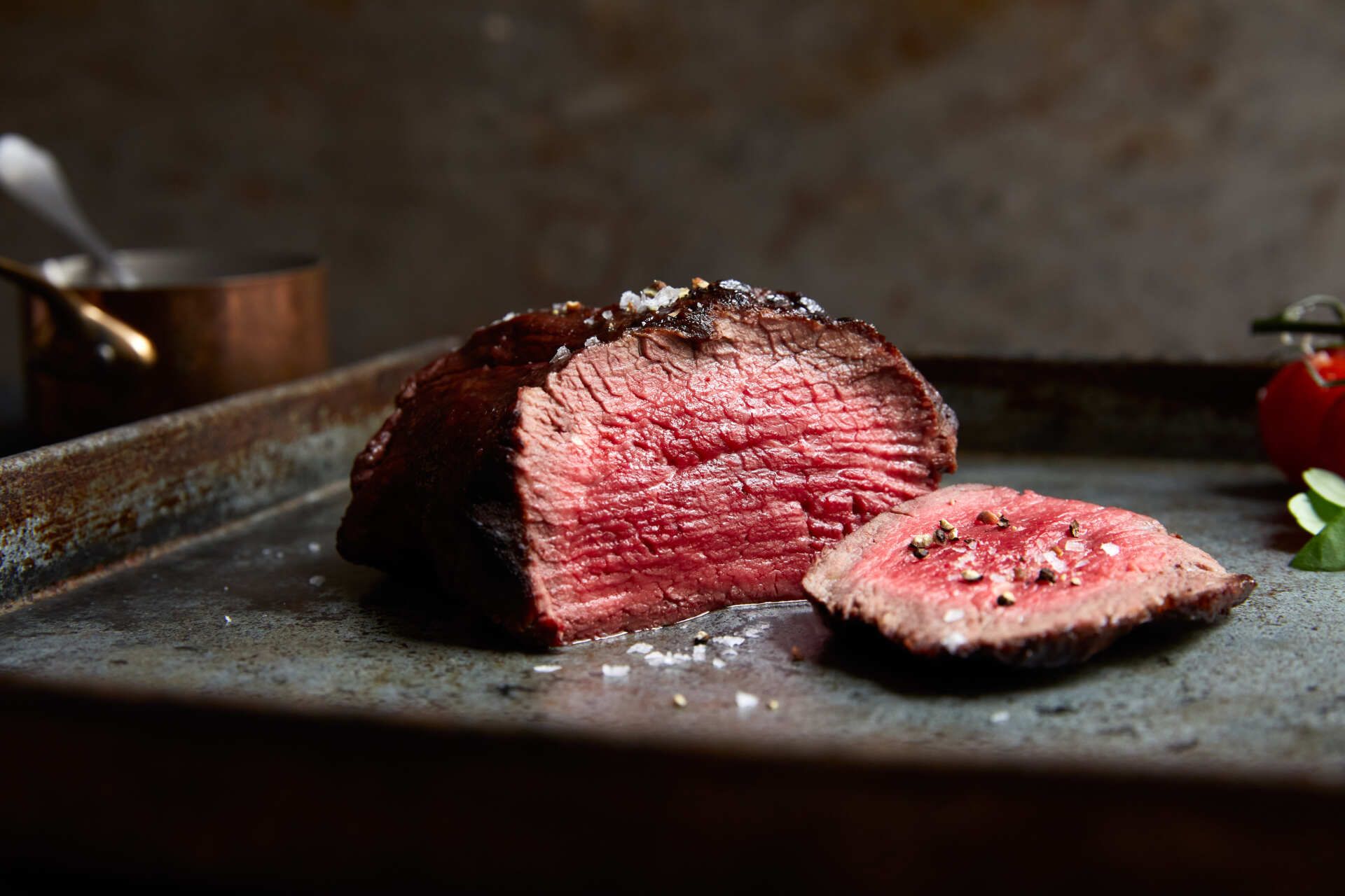 Medium-rare steak, sliced, on a metal tray, with coarse salt, against a rustic background.