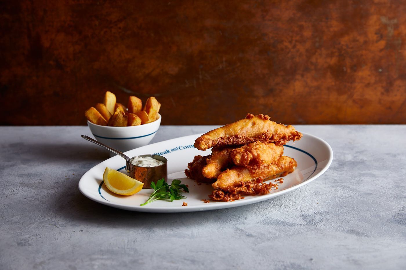 Plate of fish, tartar sauce and chunky chips on a light marble table.
