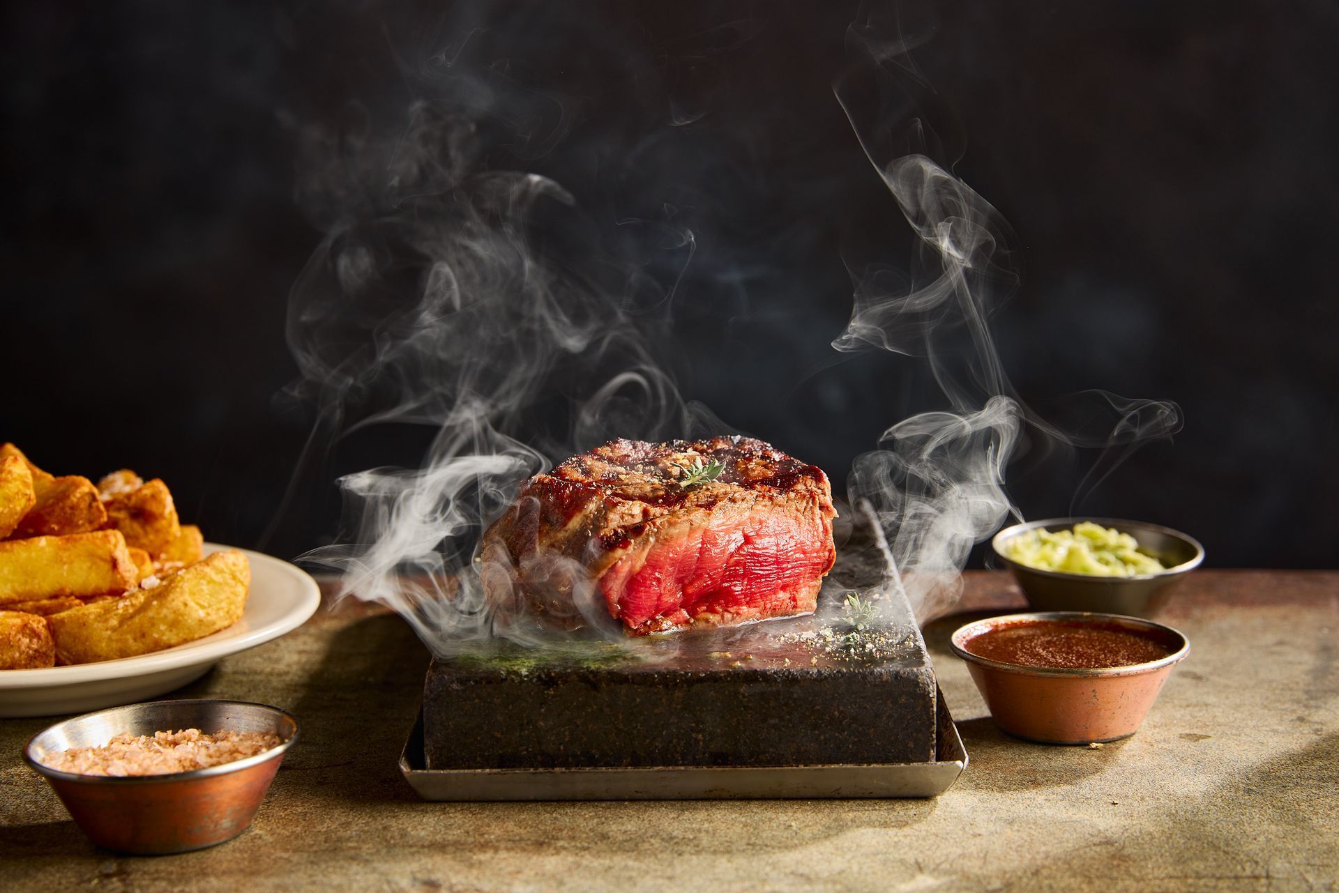 Steak on a hot stone with smoke, fries, sauces, and salt.