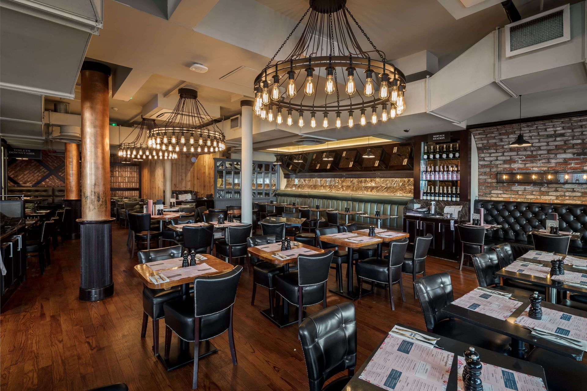 Steak and company Gloucester road Restaurant interior with tables, chairs, and ornate lighting fixtures. Dark wood floors, brick walls, and a bar area.