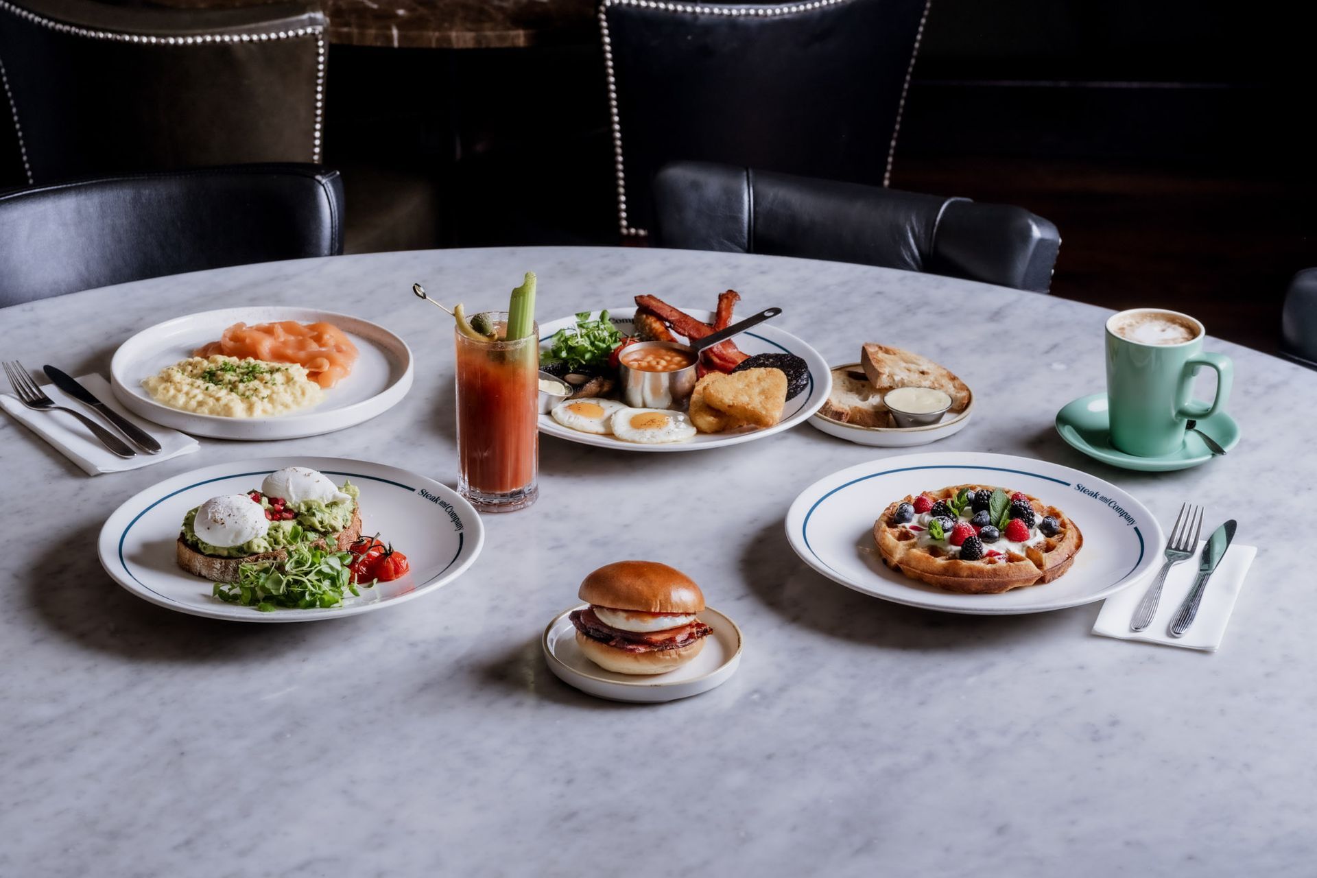 A Bloody Mary, Full English, eggs on sourdough, a waffle, scrambled eggs and a breakfast sandwich on a light marble table.