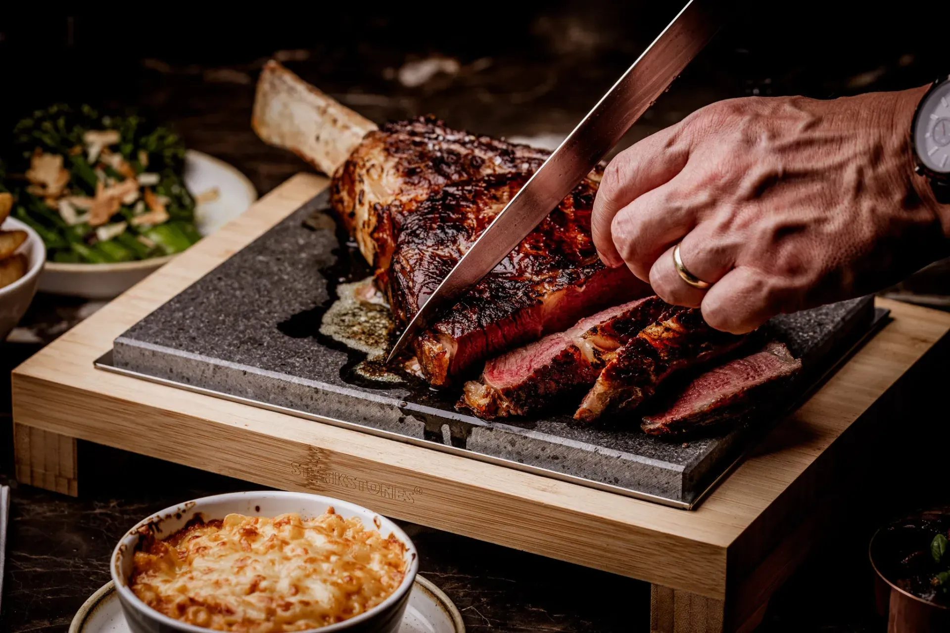 Hand slicing a large, cooked tomahawk steak on a wooden serving board, with side dishes visible.