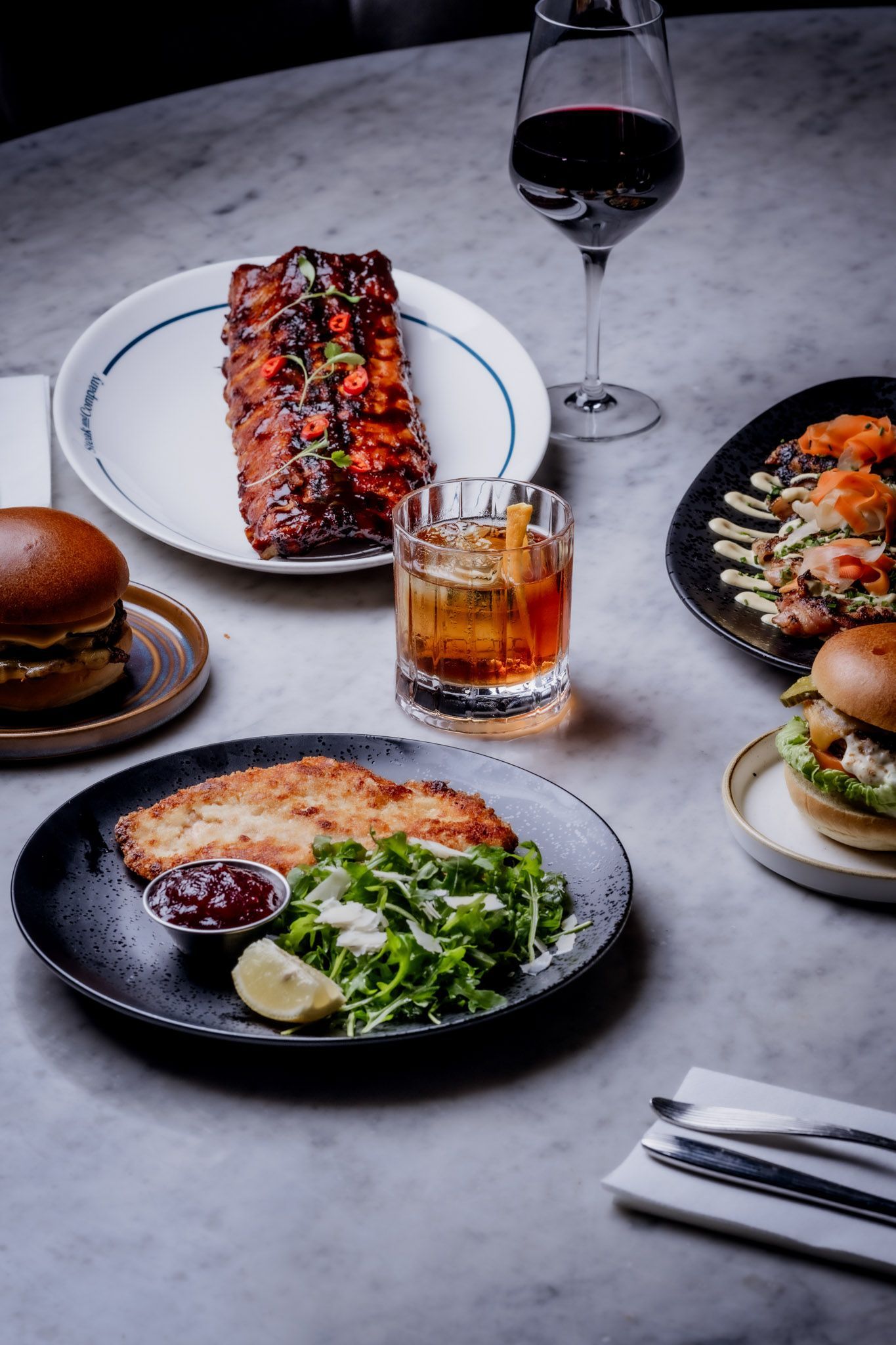 Table with various dishes: ribs, burger, fried cutlet, salad, and a cocktail, alongside silverware and a glass of wine.