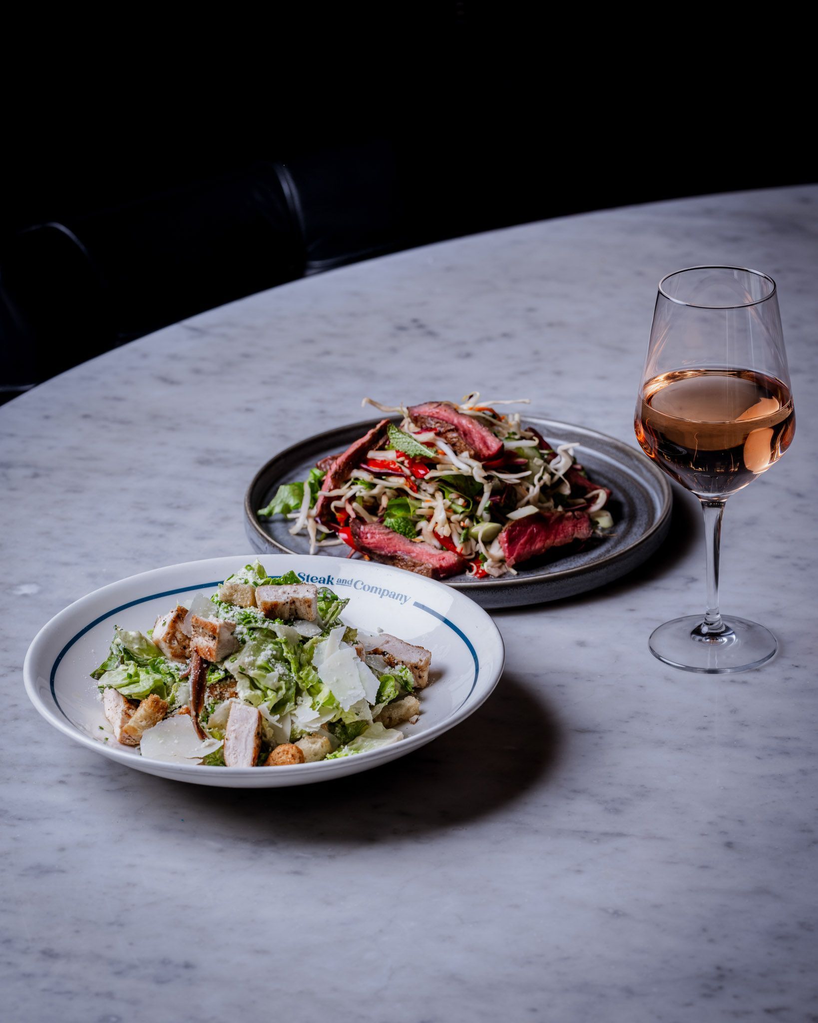 Two salads, steak and chicken Caesar, with a glass of rose wine on a marble table.
