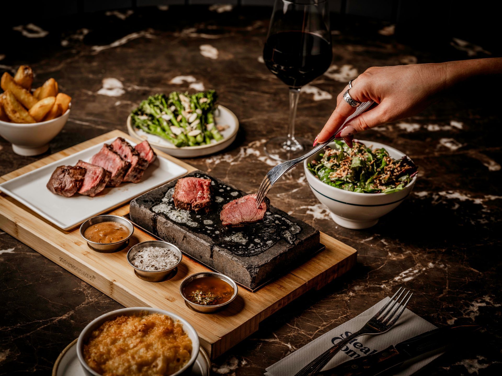 A cooked steak on a hot stone, with Chimichurri, salt and butter, on a marble table with Mac n Cheese, broccoli, a green salad and a glass of red wine on a dark marble table.