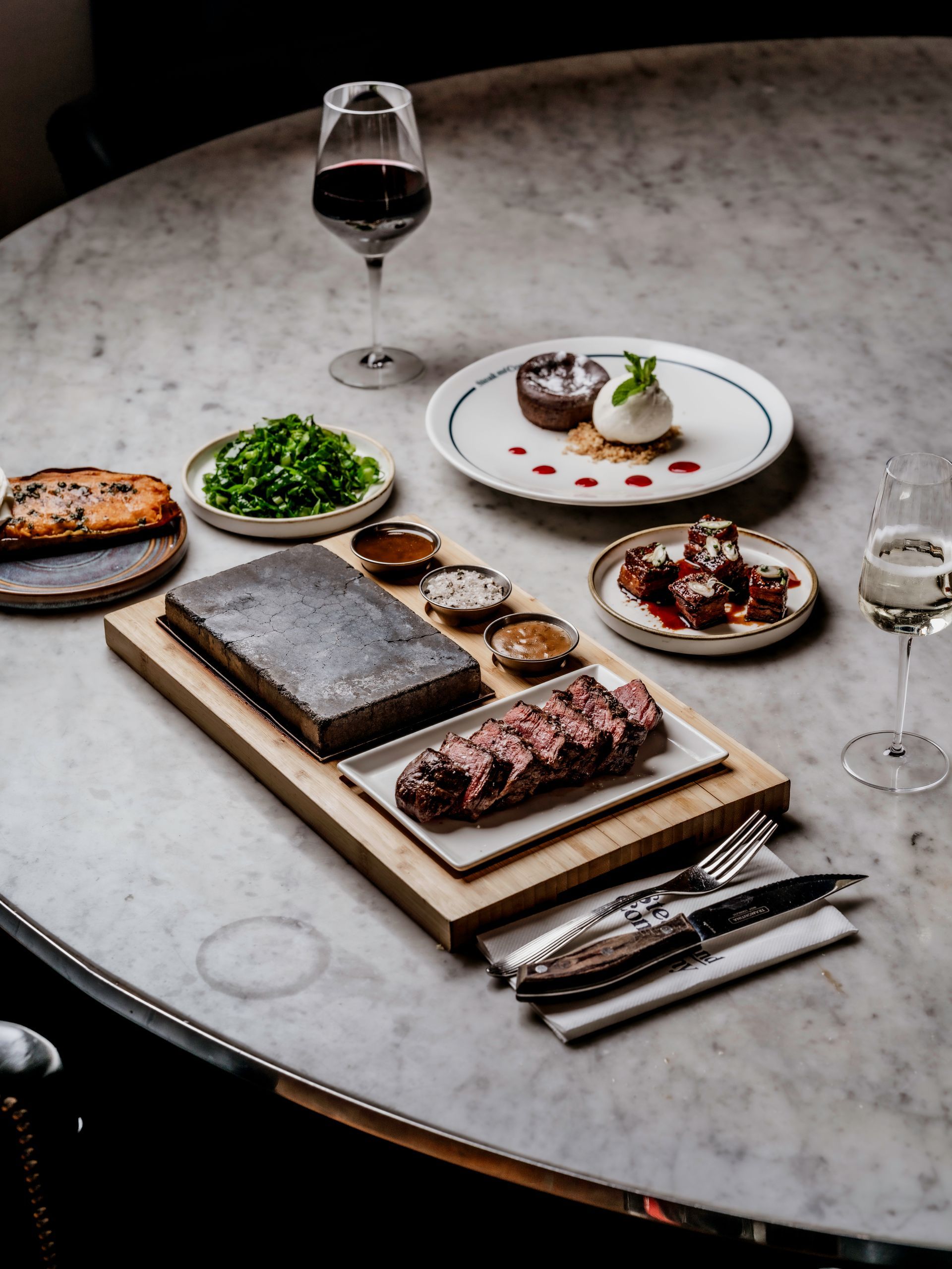 Cooked steak on a Hot Stone with Chimichrri, salt and butter, with a salad, baked potato, a tomato dish, Pork Belly Bites, a Chocolate Fondant dessert, a glass of wine and a glass of prosecco on a marble table.