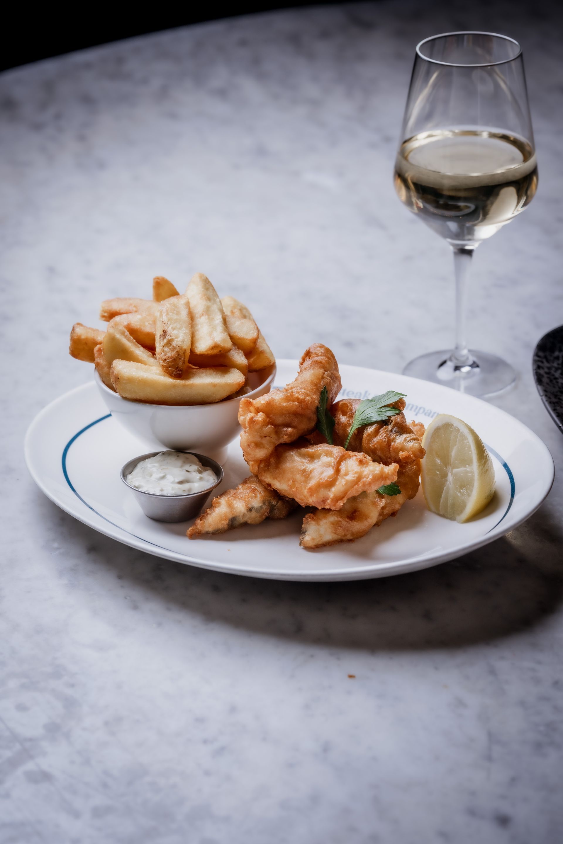 Plate of fish, tartar sauce and chunky chips on a light marble table.