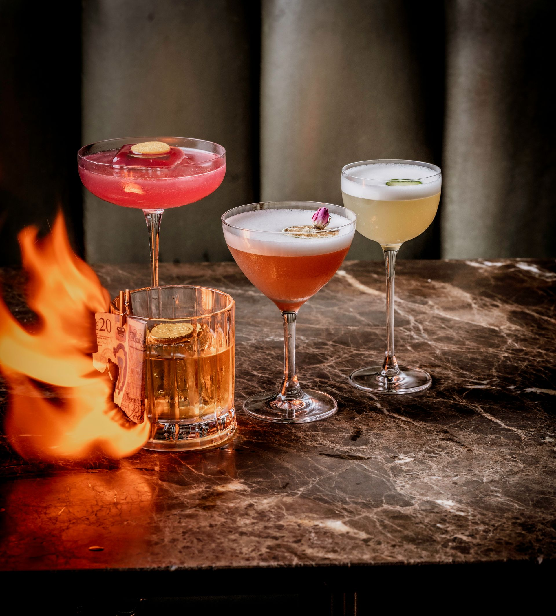 FourHappy Hour cocktails on a dark marble table, one with a note on fire.