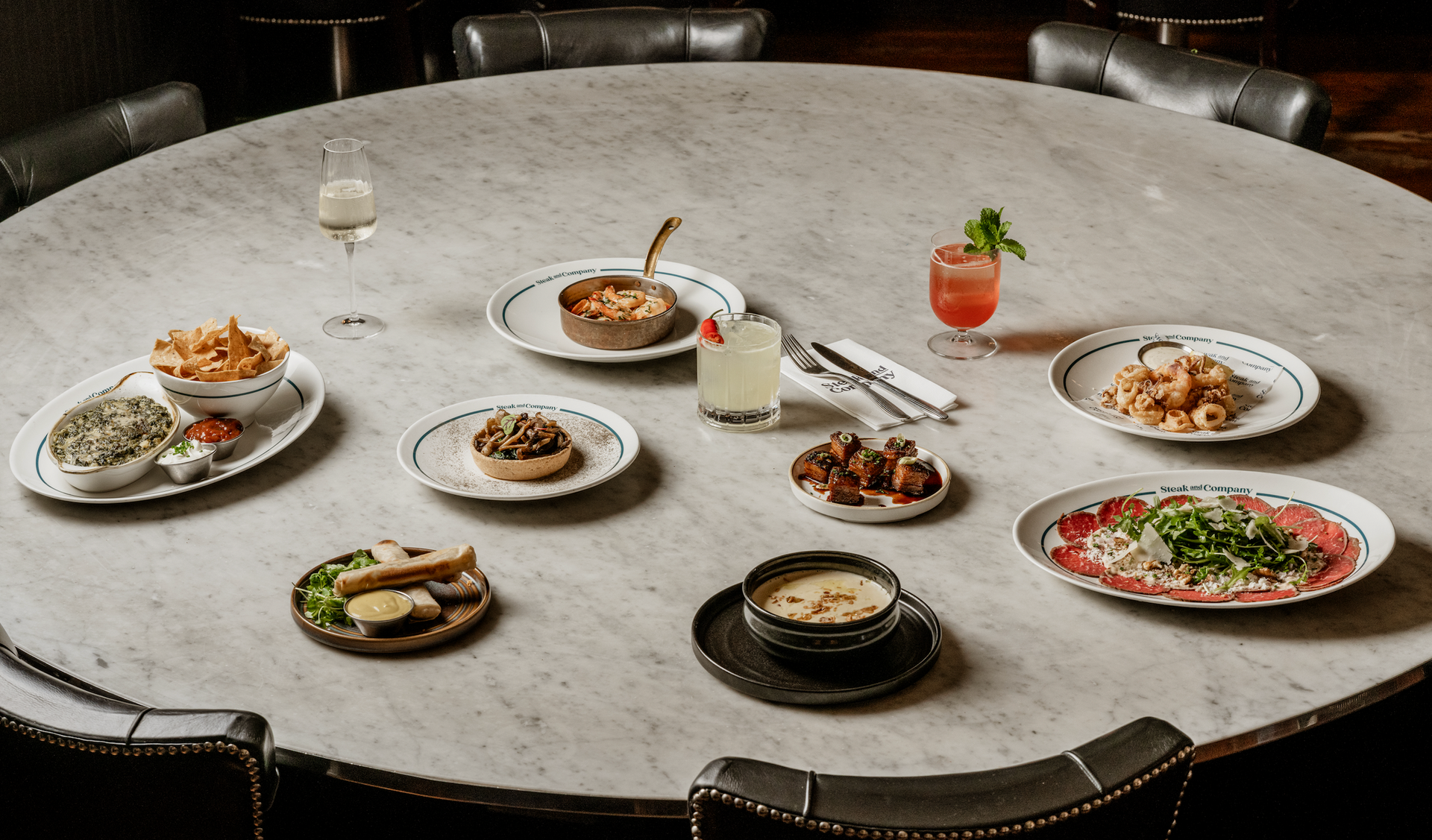 A cooked steak on a hot stone, with Chimichurri, salt and butter, on a marble table with Mac n Cheese, broccoli, a green salad and a glass of red wine on  a dark marble table.