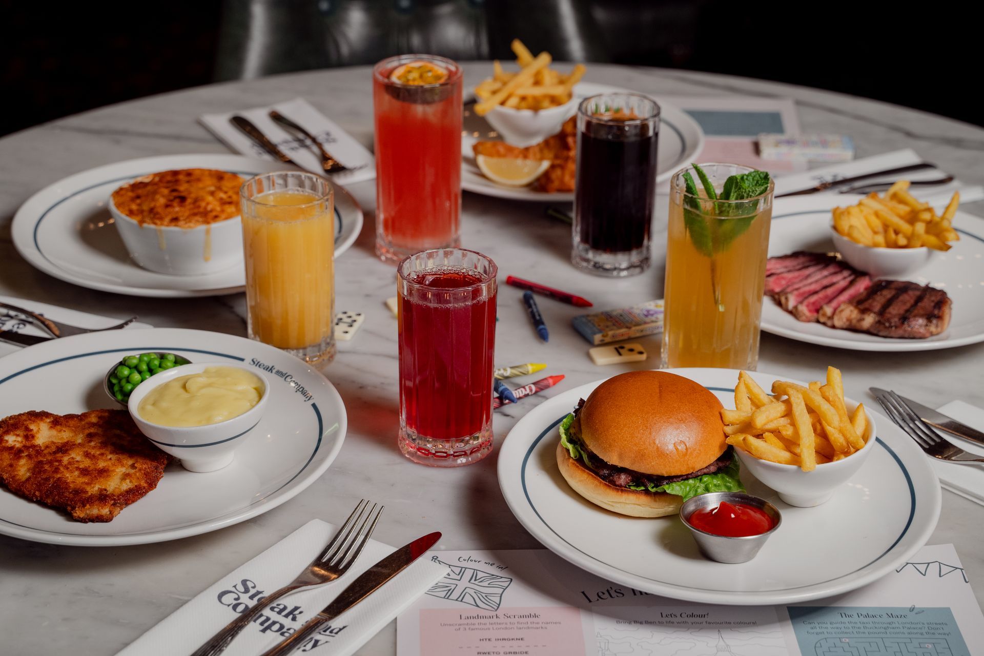 A kids burger, fries, mash, Mac n Cheese, fish and chips with soft drinks, bubble tea and crayons on a light marble table.
