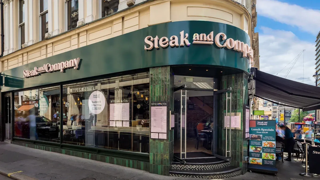 Steak and Company restaurant exterior. Green facade, large windows, outdoor seating, and a busy street scene.
