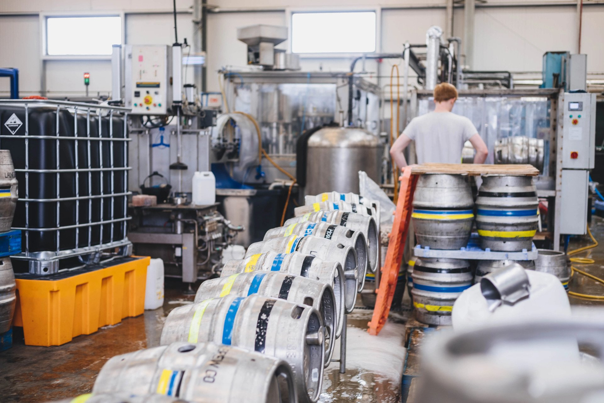 Man working with metal kegs in a factory setting, with industrial machinery and tanks in the background.