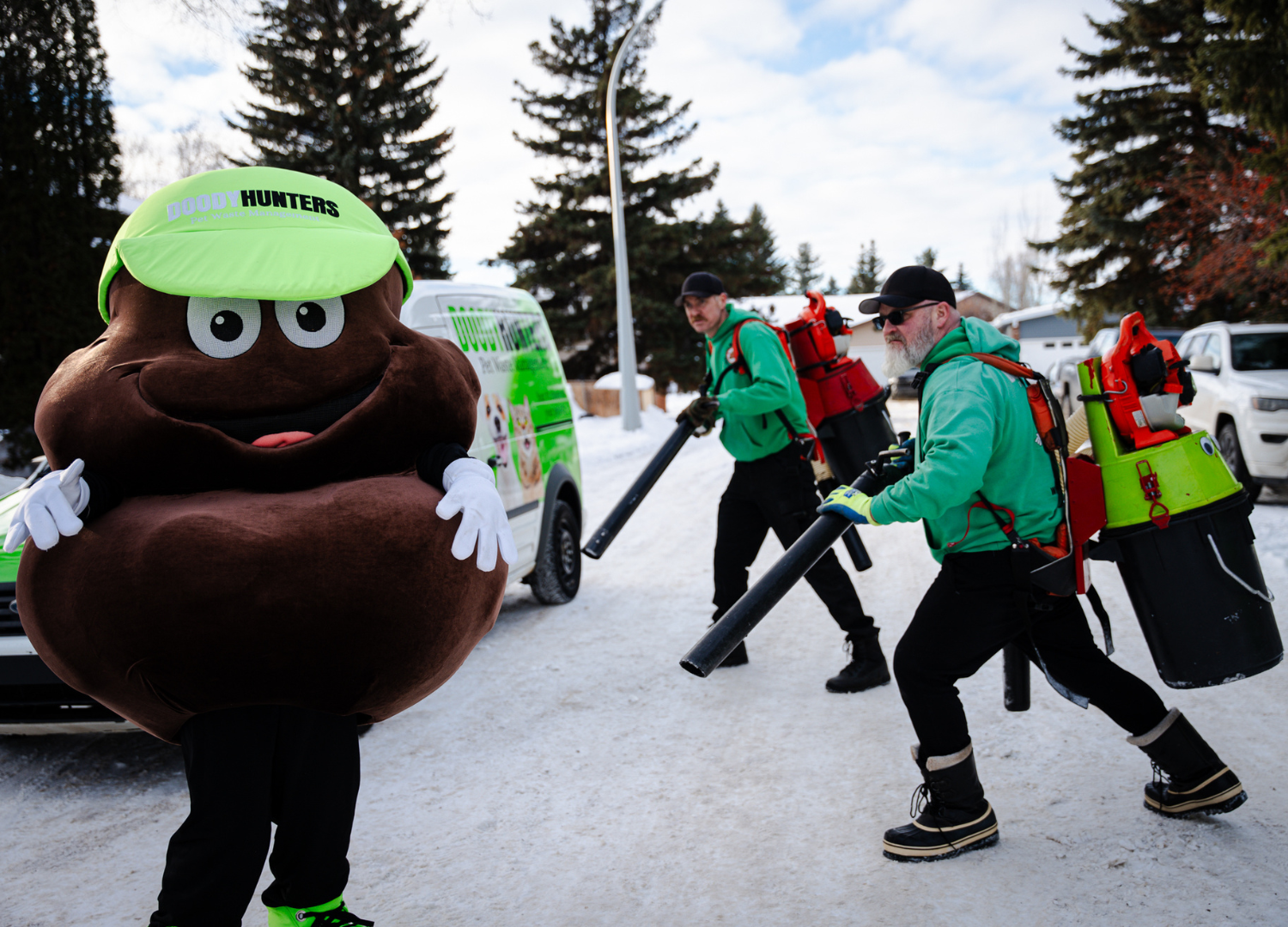 A mascot dressed as poop and two workers with leaf blowers clear snow from a street.