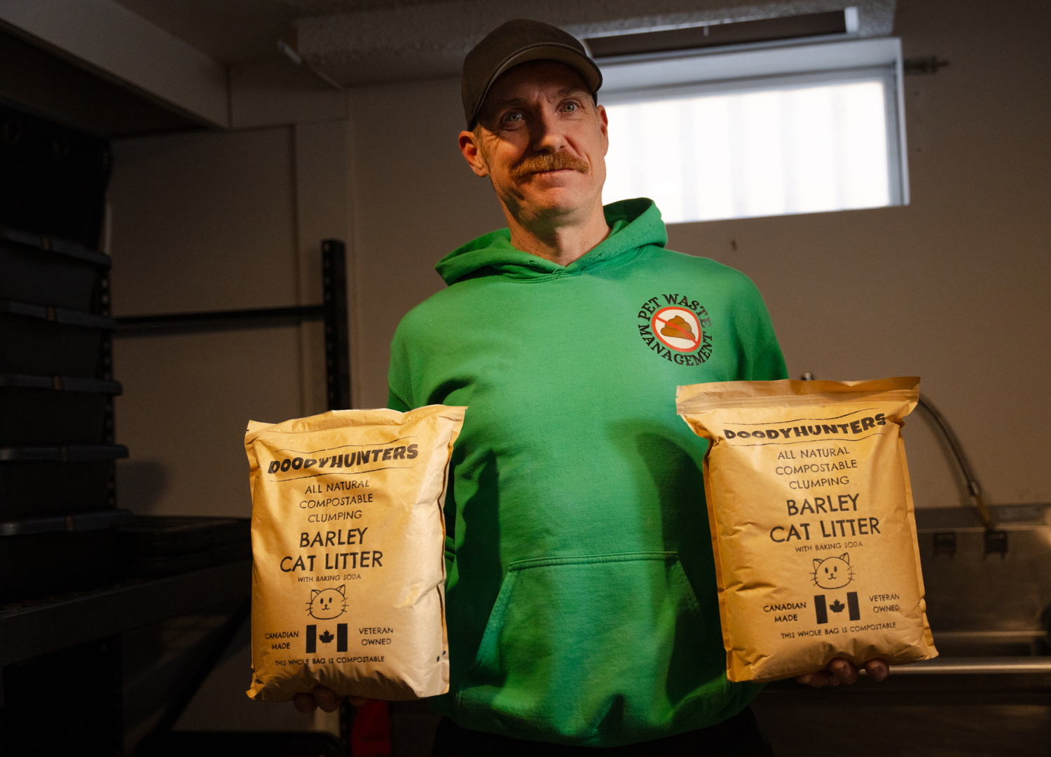 Man holding two bags of cat litter in a room. He wears a green hoodie, and a cap.